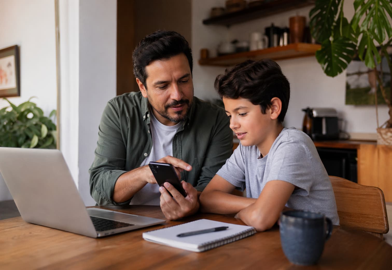 Padre e hijo sentados juntos en la mesa del comedor revisando un teléfono celular con el portátil abierto, en una conversación cercana y tranquila sobre seguridad digital familiar.