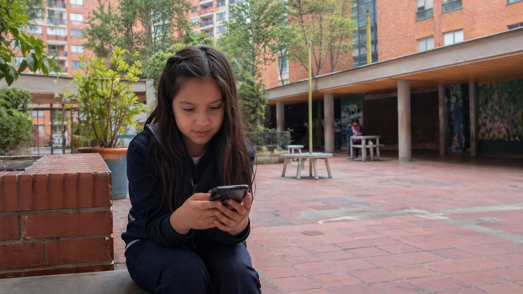Niña con uniforme escolar sentada sola en el patio del colegio, mirando su celular. Imagen que ilustra la vulnerabilidad de menores que pasan tiempo solos en entornos digitales sin acompañamiento adulto.
